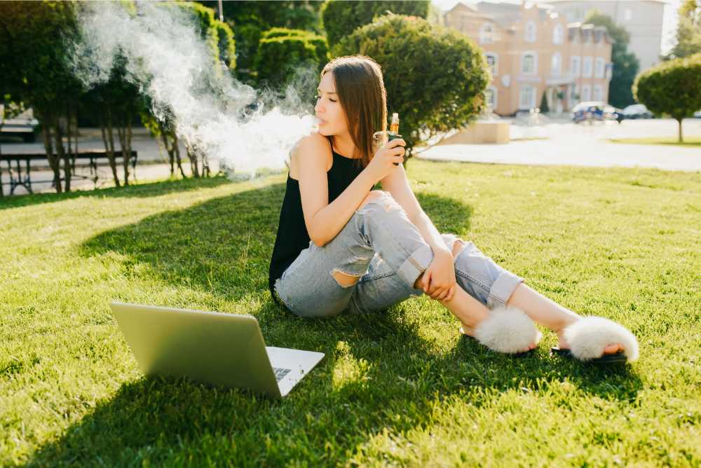 woman witting on grass facing laptop vaping